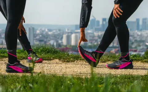 Zwei Läuferinnen dehnen sich auf einem Feldweg mit Blick auf eine Stadt. Sie tragen schwarze Laufhosen und auffällige pink-schwarze Laufschuhe. Ein Zeichen für den Wiedereinstieg ins joggen nach Geburt.