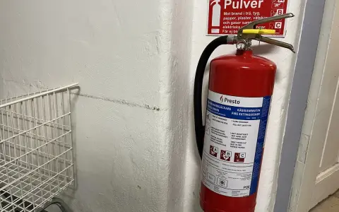 Röd brandsläckare hänger på en vit vägg, redo att användas om brandlarm ljuder. Bredvid en vit metallkorg.