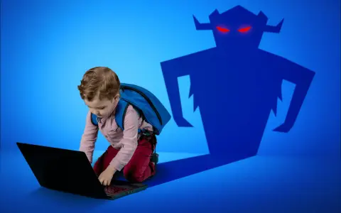 Chłopiec z plecakiem pochyla się nad laptopem, a za nim czai się złowrogi cień potwora. Zagrożenia czyhające na dzieci w internecie.
