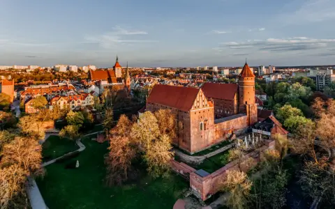 Zachód słońca nad zamkami krzyżackimi na Mazurach. Potężne ceglane mury i wieże w blasku złota, otoczone zielenią i miastem.