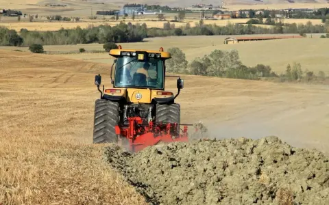 Trattore giallo in azione per l'aratura del terreno, sollevando terra e polvere in un paesaggio rurale collinare.