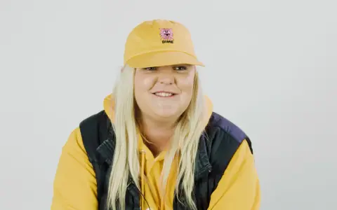 Frau mit gelber Kappe und Kapuzenpullover lächelt.