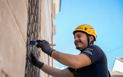 Uomo con casco giallo dipinge una grata metallica con un pennello blu. Inquadratura dal basso, cielo azzurro sullo sfondo.