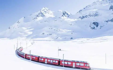 A bright red St. Moritz train winds through a pristine, snow-covered mountain landscape under a clear blue sky.