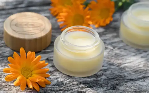 Ringelblumensalbe gegen Falten: Natürliche Pflege für glatte Haut. Kleine Gläser mit Salbe und leuchtend orangefarbene Ringelblumenblüten auf Holz.