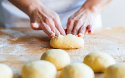 Hände formen einen **homogenen Teig** auf einer bemehlten Holzoberfläche. Mehrere Teiglinge liegen im Vordergrund.