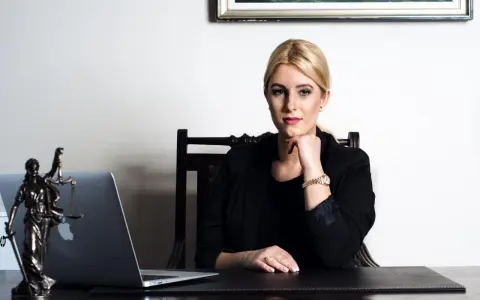 Kobieta w czarnej marynarce siedzi przy biurku z laptopem i figurką Temidy.