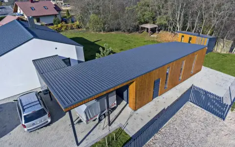 Nowoczesny budynek z drewnianą elewacją i ciemnym, falistym dachem jednopołaciowym. Obok stoi samochód i przyczepa pod zadaszeniem.