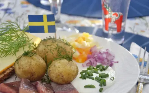 En tallrik med midsommar mat: sill, färskpotatis, ost, ägg och rödlök. En svensk flagga vajar stolt.