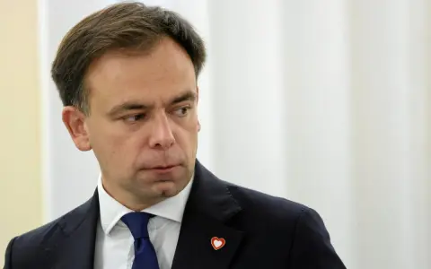 Minister finansów w granatowym garniturze i niebieskim krawacie, z przypinką w kształcie serca na klapie marynarki.