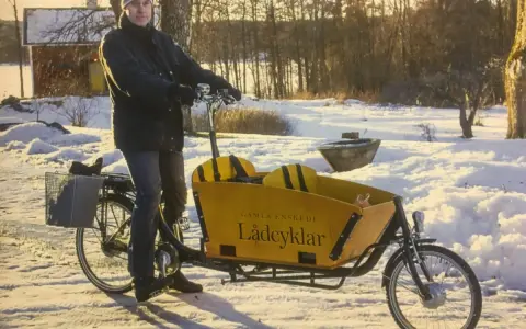 Man med mössa och jacka cyklar på vintern med en gul lastcykel i snöig miljö.