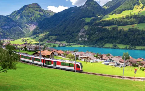 A red and white train travels through a picturesque Swiss village nestled beside a turquoise lake, with lush green mountains in the background. This is the scenic route from Lucerne to Lugano.