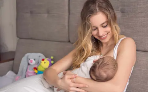 Uśmiechnięta mama karmi piersią dziecko. W tle pluszowe zabawki. Nakładki na pierś do karmienia mogą pomóc w tym intymnym momencie.