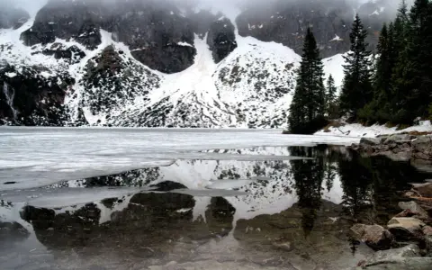 Morskie Oko zimą, zamarznięte, z odbiciem ośnieżonych gór i ciemnych drzew w przejrzystej wodzie.