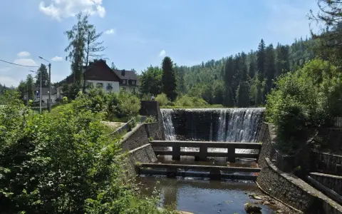 Kamienny stopień wodny na Jeziorze Czerniańskim, z wodą spływającą kaskadami. W tle zielone wzgórza i domy.
