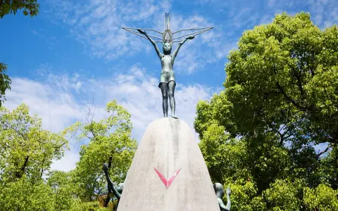 La bambina di Hiroshima, simbolo di pace, con le braccia tese verso il cielo, circondata dal verde degli alberi e dall'azzurro delle nuvole.