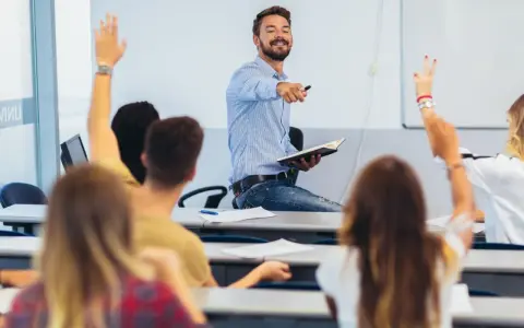 Uśmiechnięty wykładowca z książką w ręku wskazuje na studentów, którzy podnoszą ręce. Czy przed lecz stawiamy przecinek?