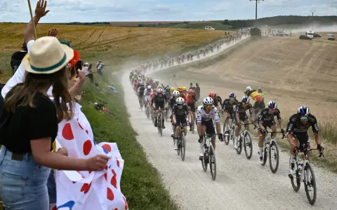 Spectators cheer as a peloton races on a dusty road during the Tour de France watch. Cyclists in colorful jerseys kick up dust in a dramatic scene.