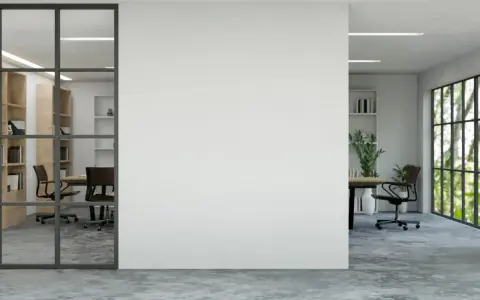 Un moderno tabique de cristal separa una sala de reuniones con estanterías de madera de una oficina con vistas a un jardín frondoso.