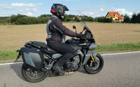 Motocyklista na szarym motocyklu Yamaha Tracer 900, z kuframi, w kasku, jedzie drogą. W tle pola i domy. Yamaha Tracer 900 dane techniczne.