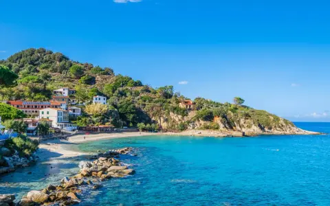 En pittoresk kustby på Elba, Italien, med vita hus som klättrar uppför en grönskande kulle. Klart turkost vatten sköljer mot en sandstrand.