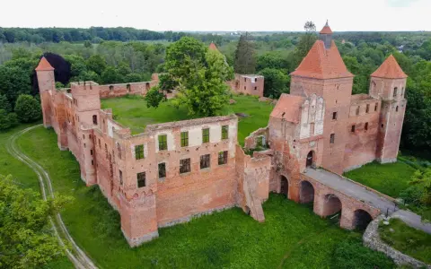 Ruiny ceglanego zamku w Szembarku otoczone zielenią. Widać most nad fosą, wieże i fragmenty murów obronnych.