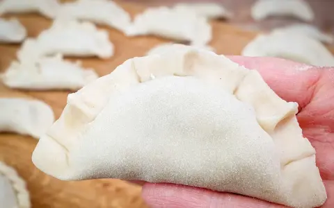 Dłoń trzyma surowe ciasto na pierogi, z innymi pierogami w tle.