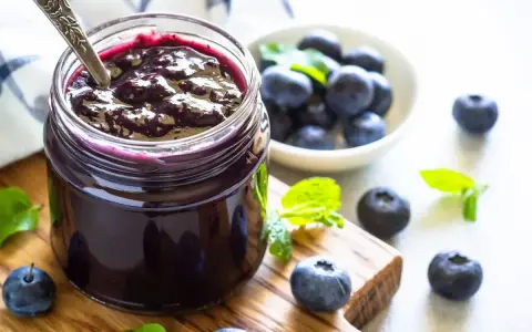 Vaso di marmellata di mirtilli fatto in casa, con mirtilli freschi e foglie di menta su un tagliere di legno.