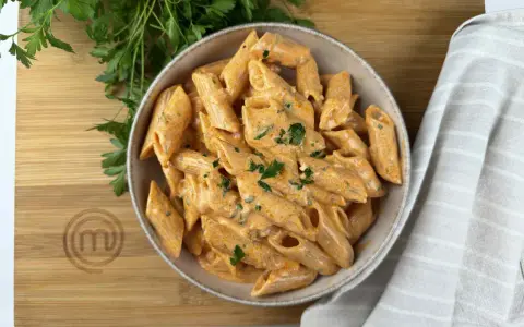 Penne alla vodka cremose e invitanti, guarnite con prezzemolo fresco, servite in una ciotola su un tagliere di legno con il logo MasterChef.