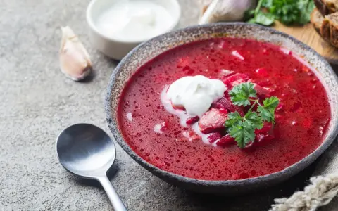 Aromaticzny barszcz czerwony domowy z łyżką śmietany i natką pietruszki, obok czosnek i chleb.