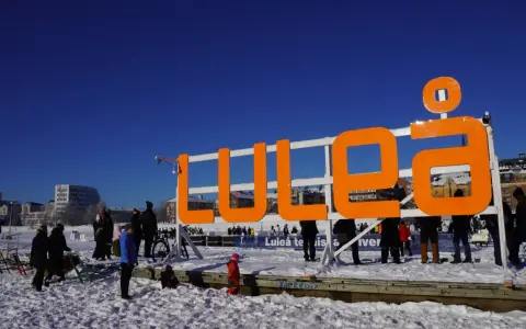 Stor orange skylt med texten "LULEÅ" i snöig miljö. Perfekt för luleå travel, en solig vinterdag.