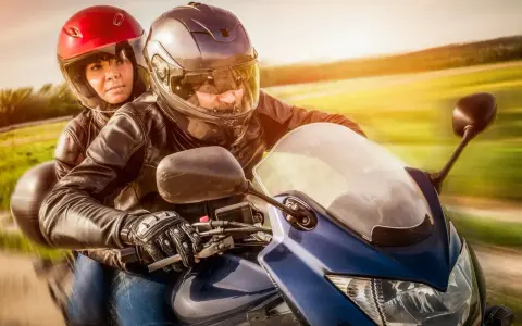 Coppia in moto, con lui alla guida e lei dietro, entrambi con caschi e giacche di pelle. Un'immagine che evoca la libertà di come guidare una moto.