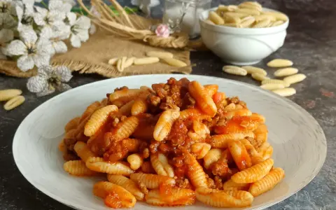 Un piatto invitante di malloreddus alla campidanese, conditi con un ricco ragù di carne. Sullo sfondo, fiori bianchi e un ciotola di pasta cruda.