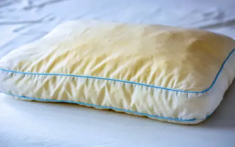 Ein vergilbtes Kissen mit blauen Nähten, das auf einem weißen Bett liegt. Gelbe Flecken deuten auf möglichen Schimmel hin.