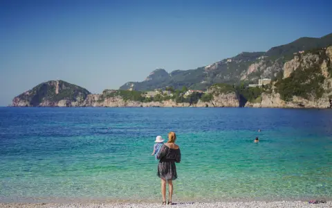 Kobieta z dzieckiem na rękach podziwia piękne Korfu plaże. Woda jest krystalicznie czysta, a w oddali widać góry.