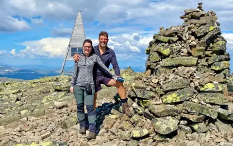 Två glada vandrare njuter av utsikten från en bergstopp under en vandring i Sverige. En stenröse och en modern träkonstruktion med solpaneler syns i bakgrunden.