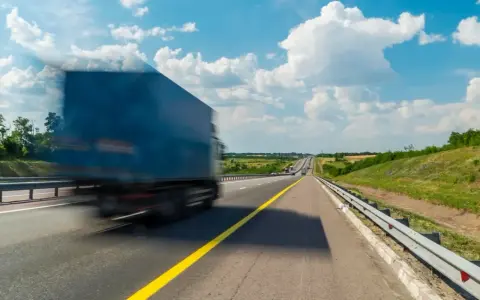 Niebieska ciężarówka w ruchu na autostradzie pod błękitnym niebem z chmurami.