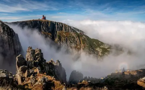 Schronisko na szczycie góry, otoczone mgłą i skalistymi zboczami. Widok na śnieżne kotły i tęczę.