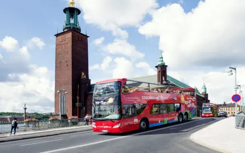 Röd dubbeldäckare med "Hop-on Hop-off Stockholm Buses" på väg förbi Stadshuset. Perfekt för stockholm turistinformation.