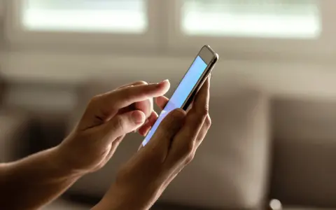 Dłonie trzymają smartfon, na ekranie którego wyświetla się niebieski kolor. Zastanawiasz się, jak sprawdzić ile lat ma telefon?