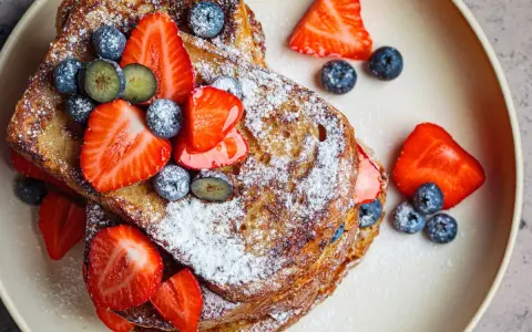 En hög med gyllene french toast, toppad med färska jordgubbar, blåbär och pudersocker. Ett perfekt recept för en lyxig frukost.