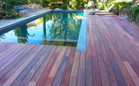 Terrasse en padouk vieilissant, reflétant les arbres dans la piscine. Un transat attend au soleil.