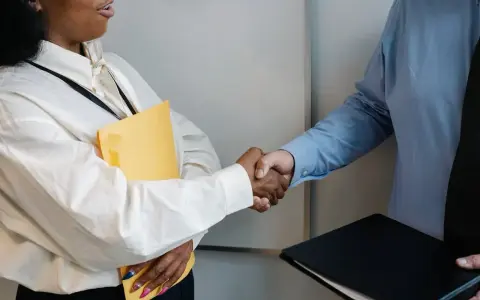 Uścisk dłoni po rozmowie o przedłużenie umowy o pracę. Kobieta trzyma żółtą teczkę, mężczyzna czarną.