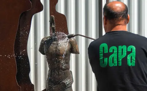 Hombre con camiseta negra "Capa" limpia una estatua de bronce con agua a presión.