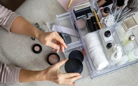 Hände sortieren Kosmetik in einem transparenten Organizer mit Pinseln, Puderdosen und Cremes.