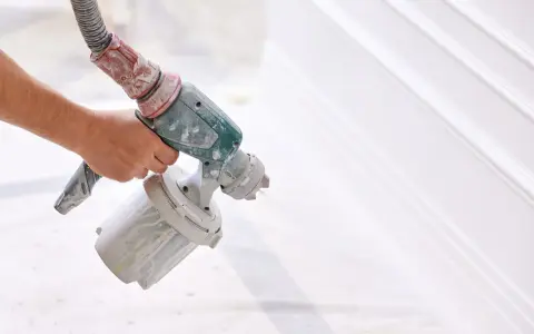 Mano che tiene una pistola a spruzzo per verniciare un cancello bianco, creando un effetto liscio e uniforme.
