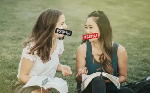 Two women with censored mouths discuss if cursing is a sin while reading books on the grass.
