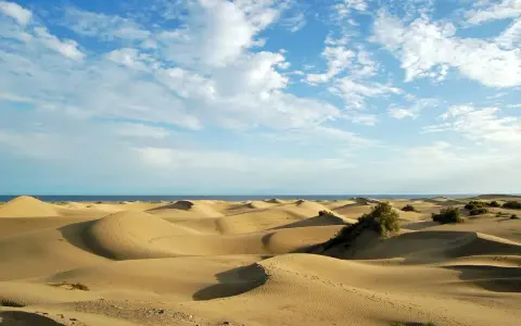 Paisaje de dunas doradas en España, con el mar azul al fondo y un cielo despejado con nubes blancas.