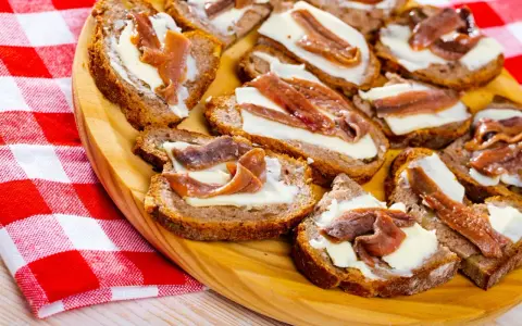 Na drewnianej desce ułożone są kromki chleba z masłem i anchois. To pyszne, proste przekąski.