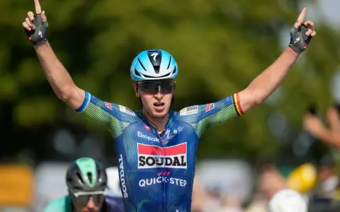 Tim Merlier jubelt mit erhobenen Armen und zeigt den Sieg. Er trägt ein blau-grünes Radtrikot mit Sponsorenlogos.